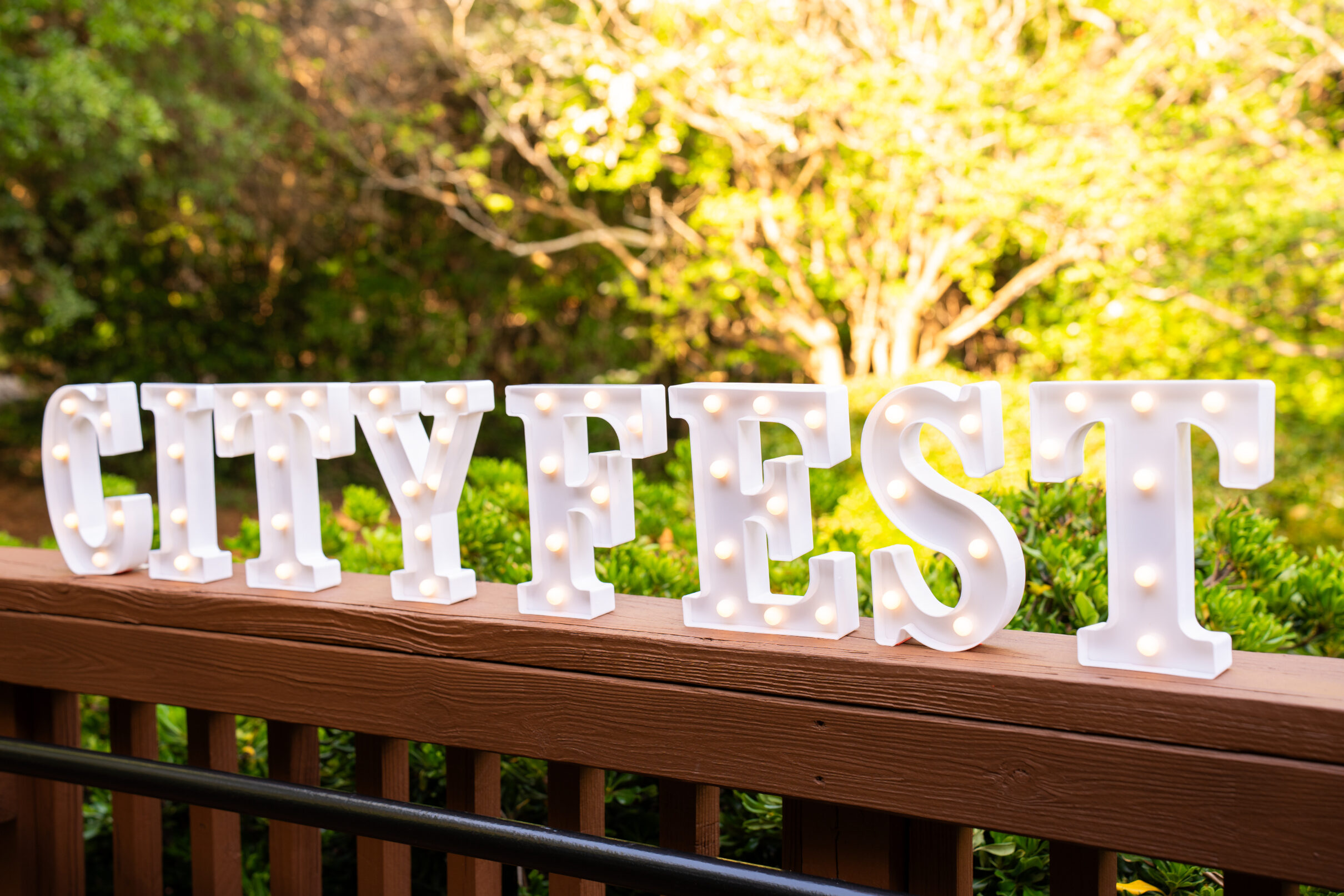White marquee-style letters spelling “CITY FEST” with small lights, displayed on a wooden railing with greenery in the background.