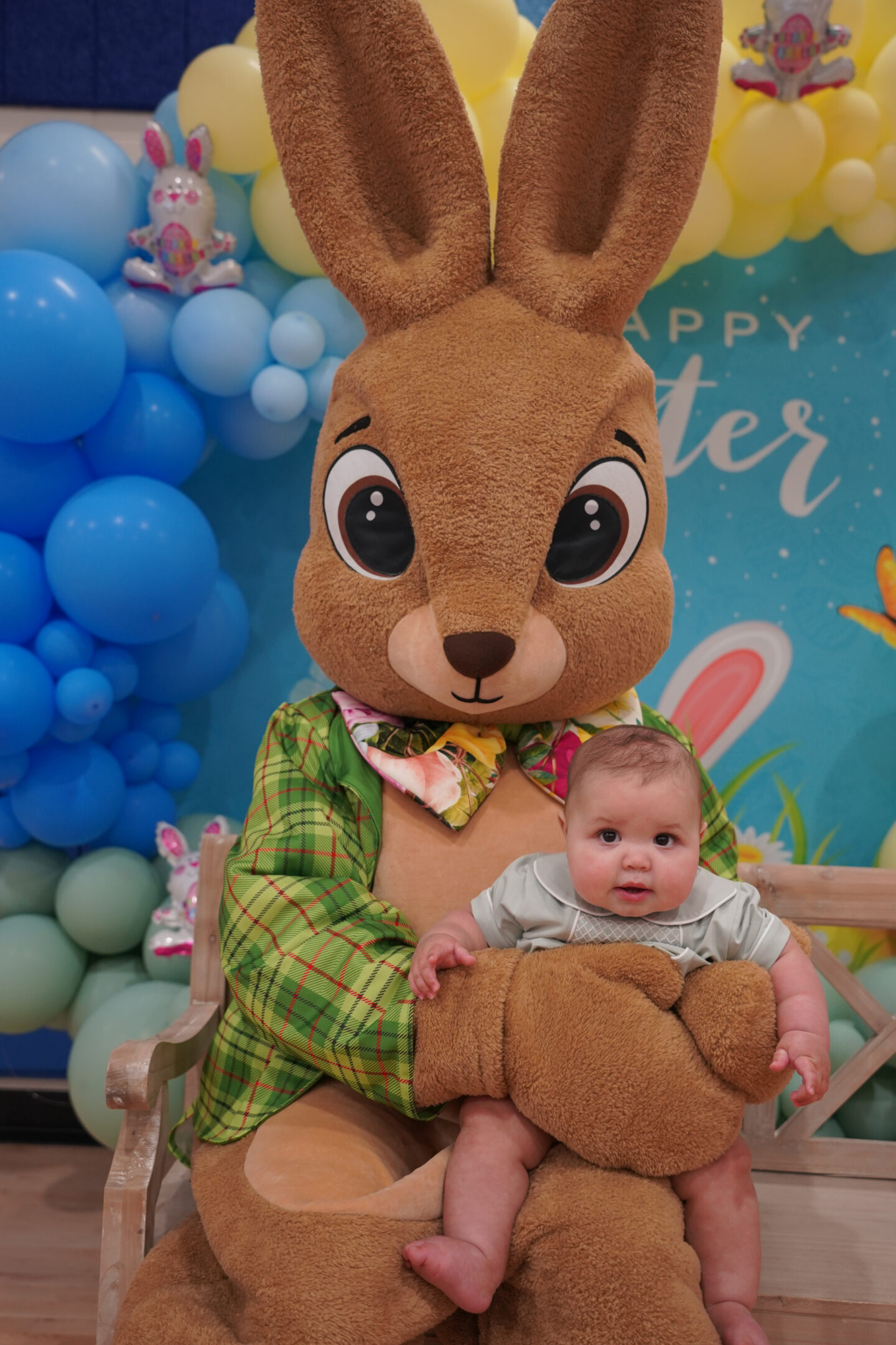 A child being held by an Easter bunny while seated on a bench in front of a colorful, assorted baloon backdrop.