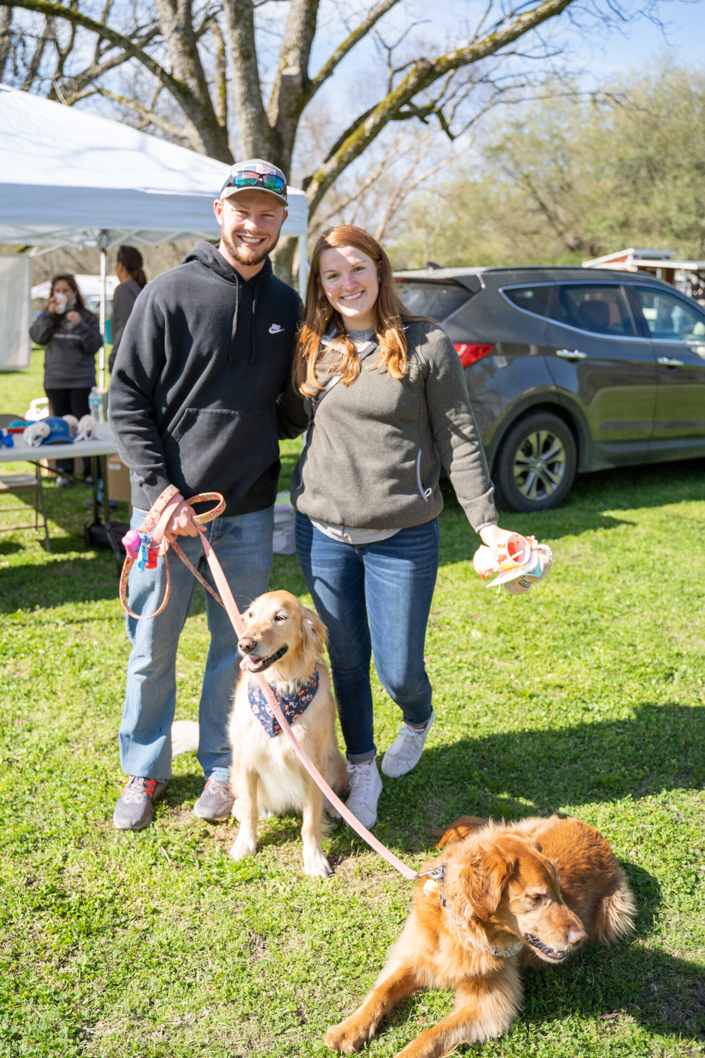 Couple smiling and posing with two dogs on leashes at an outdoor event, with tents, people, and parked cars in the background.