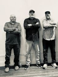 Black and white image of three men standing indoors with their arms crossed while staring at the camera.
