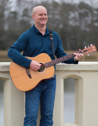 Man stands outdoors holding guitar and smiling. 