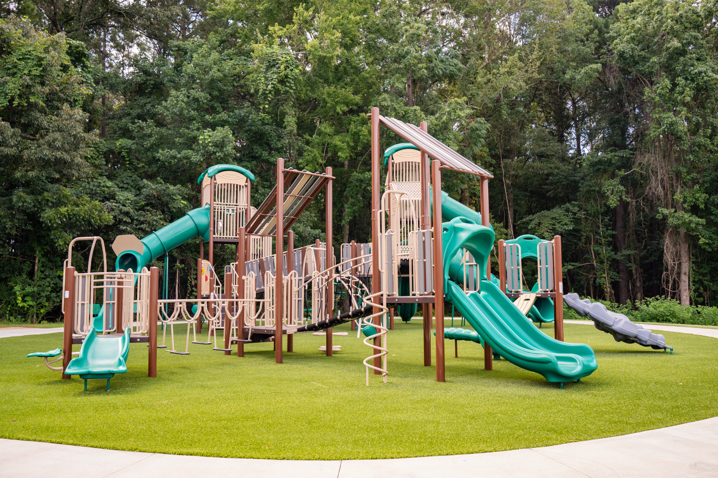 Playground with slides, climbing structures, and bridges set on a grassy area, surrounded by trees.