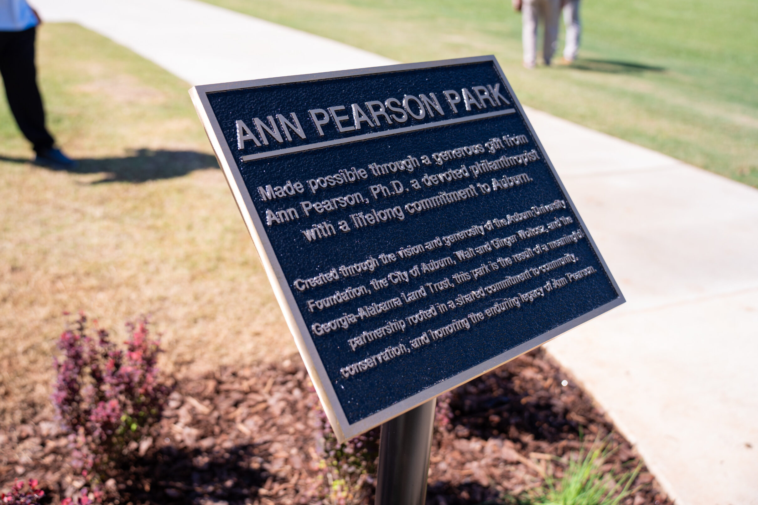 Plaque at Ann Pearson Park with engraved text recognizing the park’s namesake and contributors, set beside a sidewalk and landscaped area.
