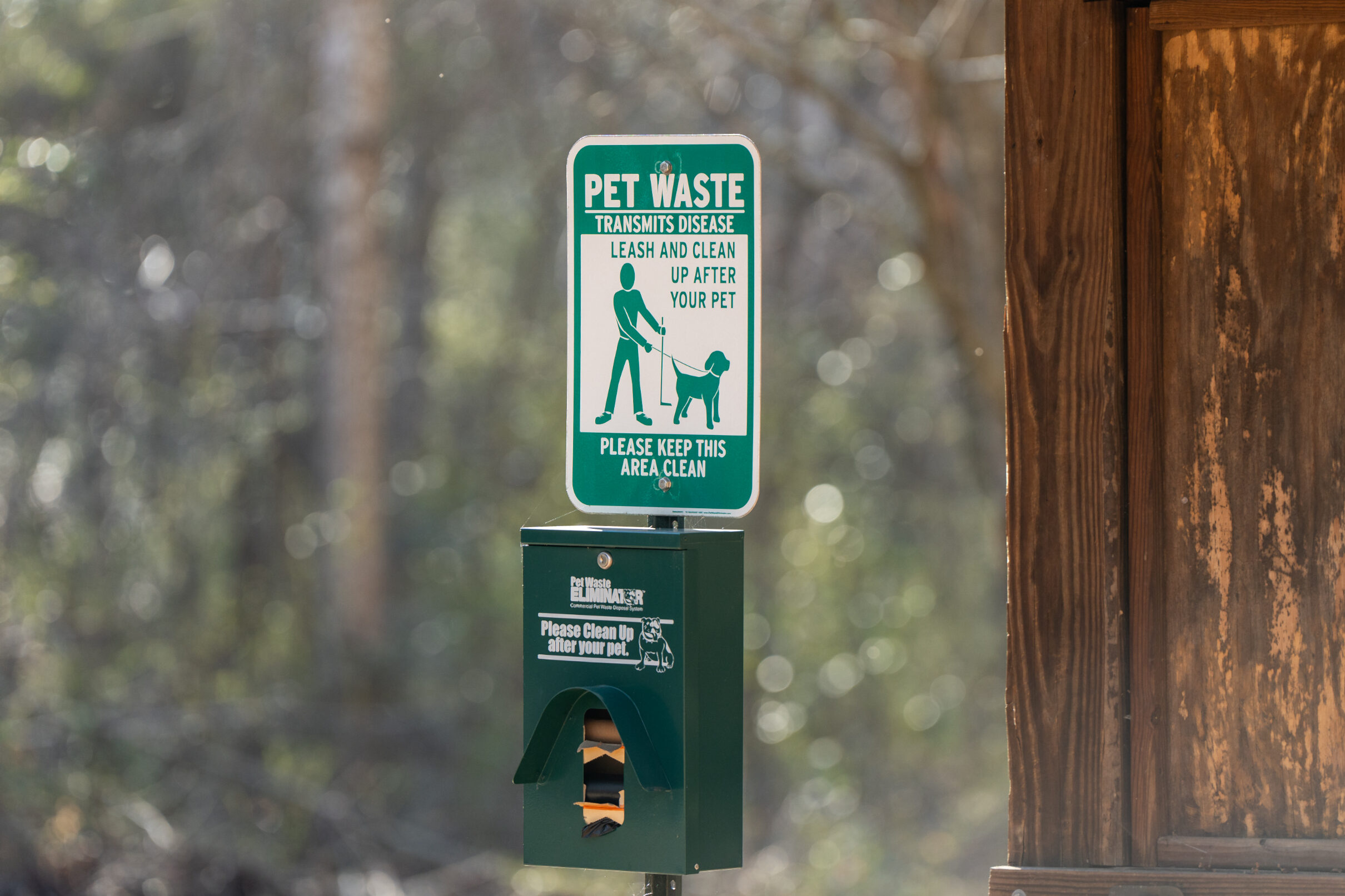 Pet waste station with sign reminding visitors to leash pets and clean up waste, including a dispenser for bags in a wooded park setting.