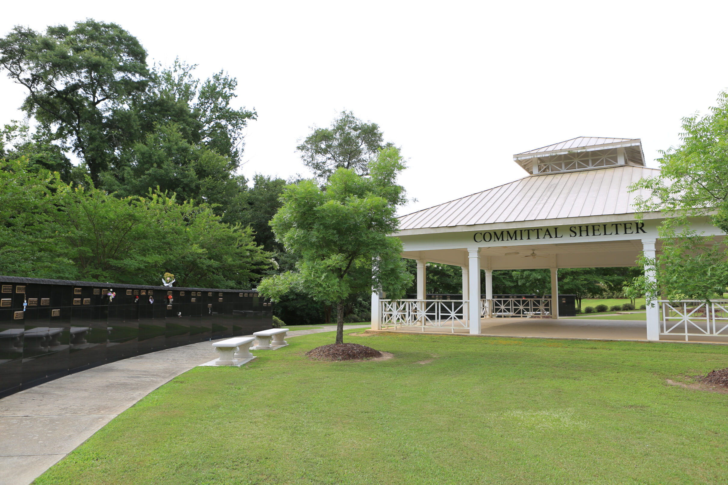 Open-air committal shelter at a cemetery with a covered pavilion, surrounding columbarium walls, benches, and landscaped grounds.
