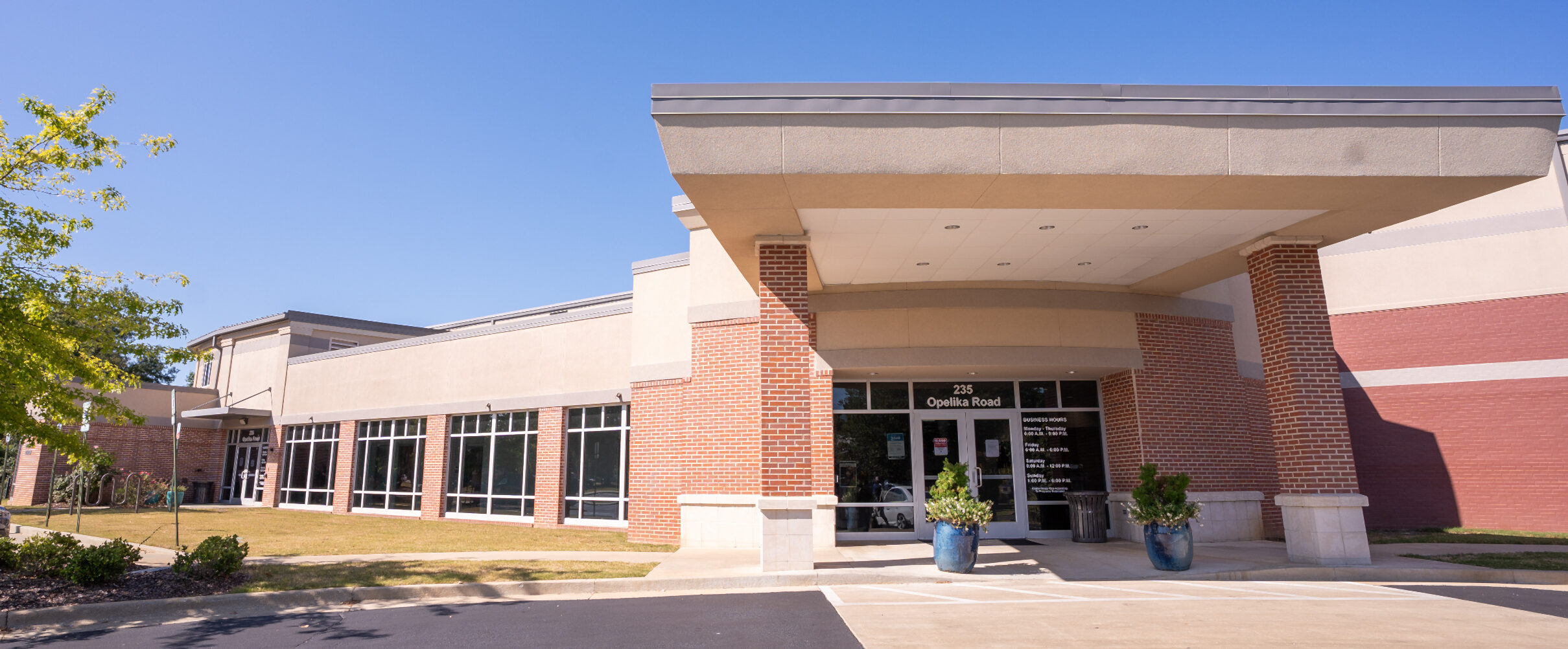 Main entrance of a recreation center featuring a covered drop-off area supported by brick columns, with accessible glass double doors, adjacent windows, and a paved walkway leading from the parking area.