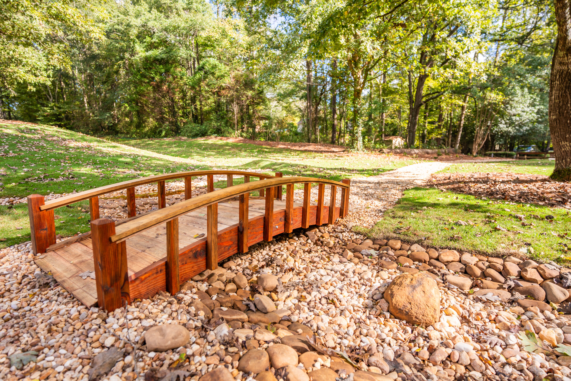 Small wooden footbridge over a dry creek bed in a landscaped park with trees and a walking path.
