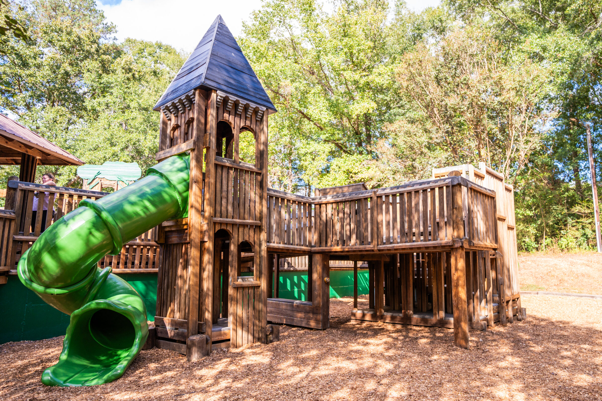 Wooden playground structure with a tower, elevated platforms, and a green spiral slide, set in a wooded park area.