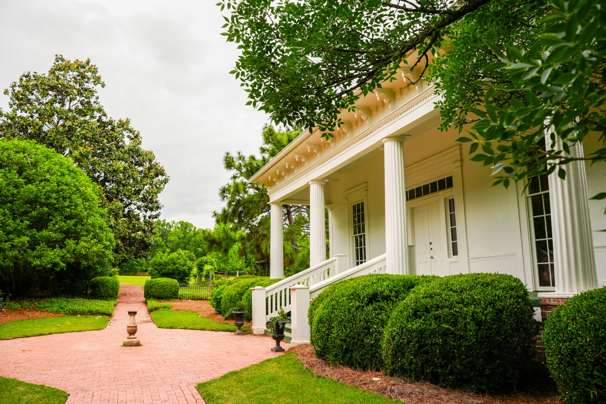 Brick pathway leading to a white columned building with landscaped hedges and trees in a park setting.