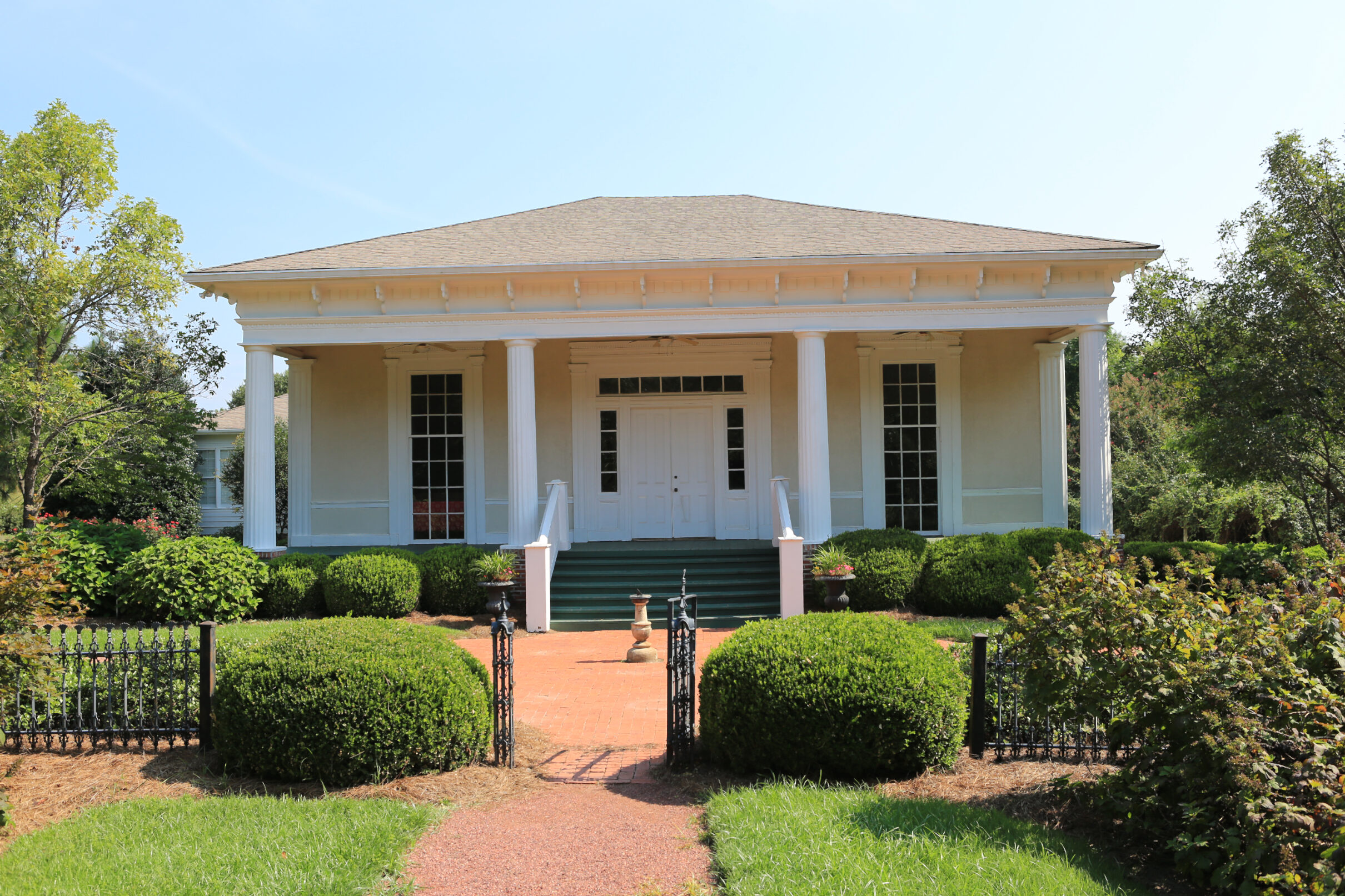 White columned building set in a landscaped park, with a brick path, bench, and surrounding greenery.