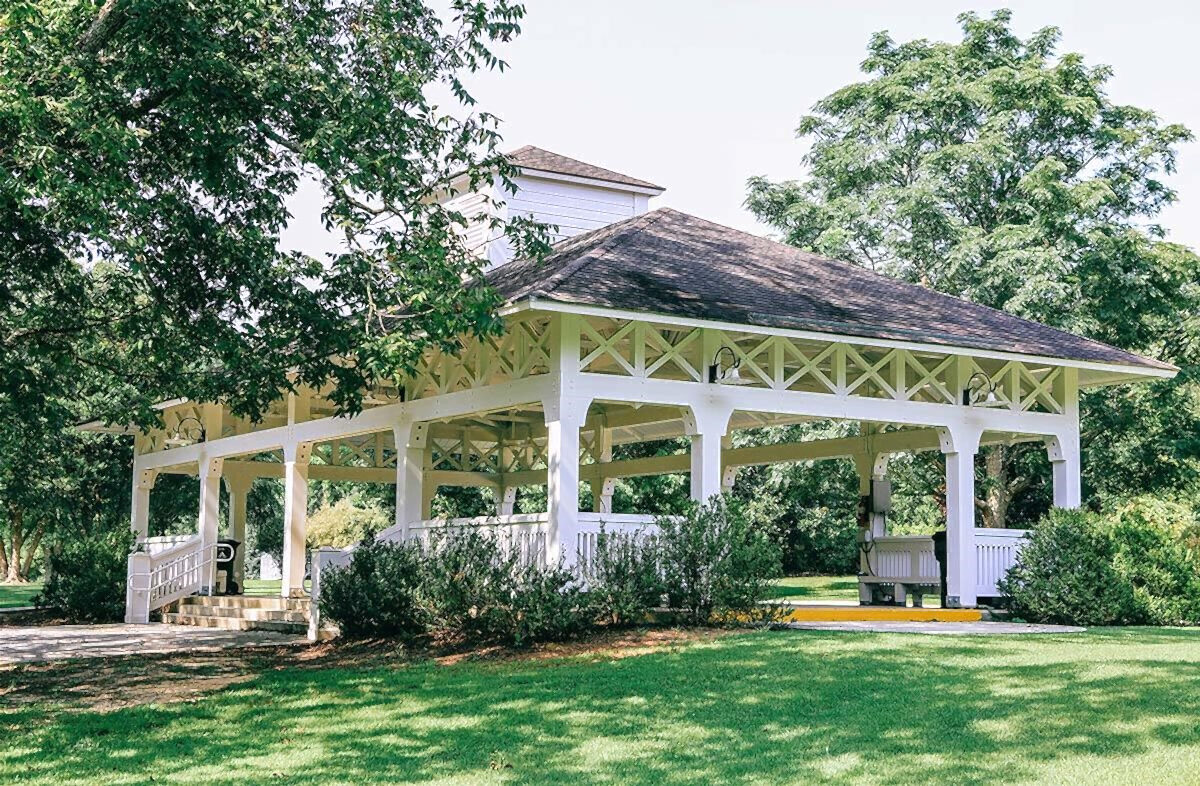 Open-air park pavilion with white columns and a shingled roof, surrounded by trees and landscaped greenery.