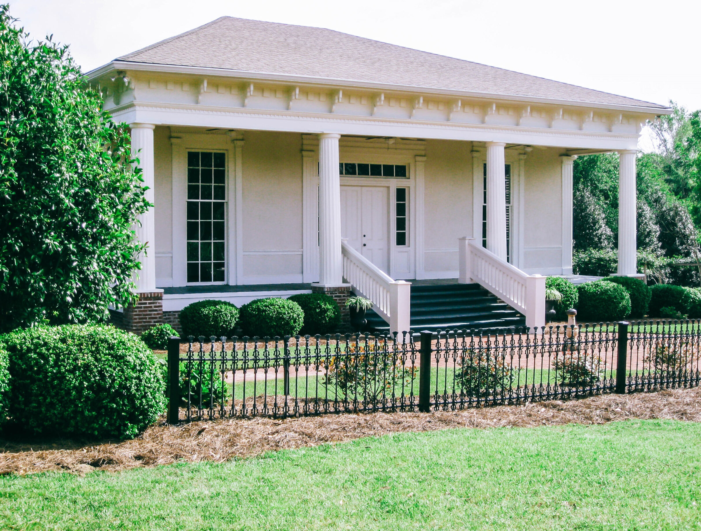 White columned building with a front porch and steps, surrounded by landscaped hedges and a decorative metal fence.