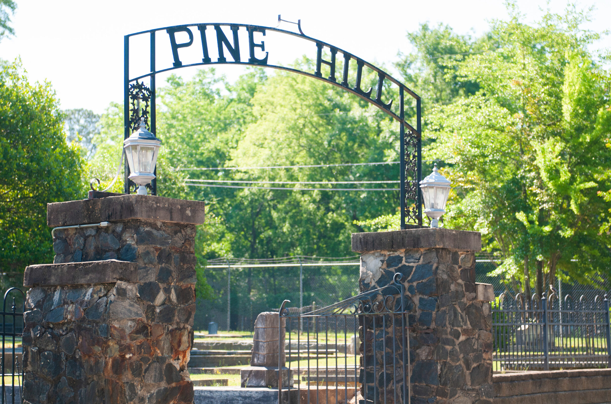 Stone entrance pillars with lanterns and an iron arch reading “Pine Hill,” marking the entrance to a cemetery with fencing and trees beyond.