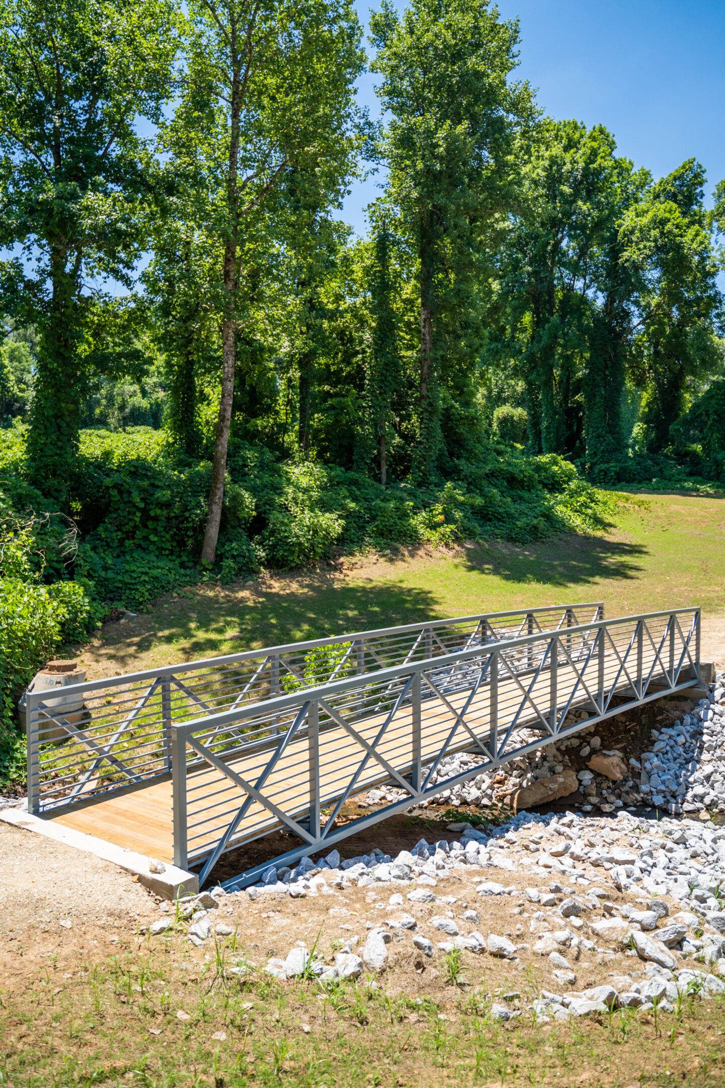 Metal pedestrian bridge crossing a small rocky creek, with a wooded area and grassy bank nearby.