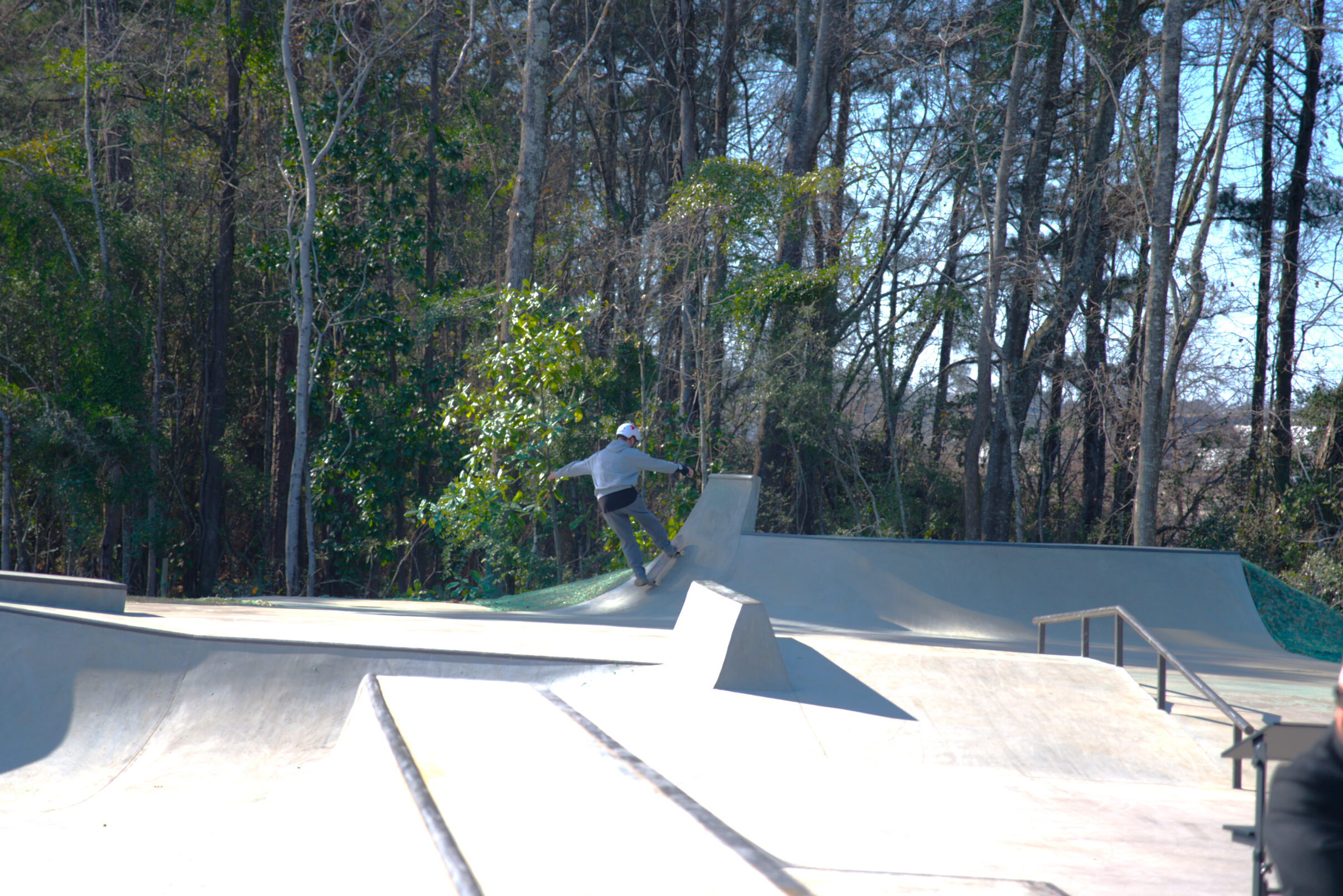 Person skateboarding on a ramp at a concrete skate park with rails and wooded surroundings.
