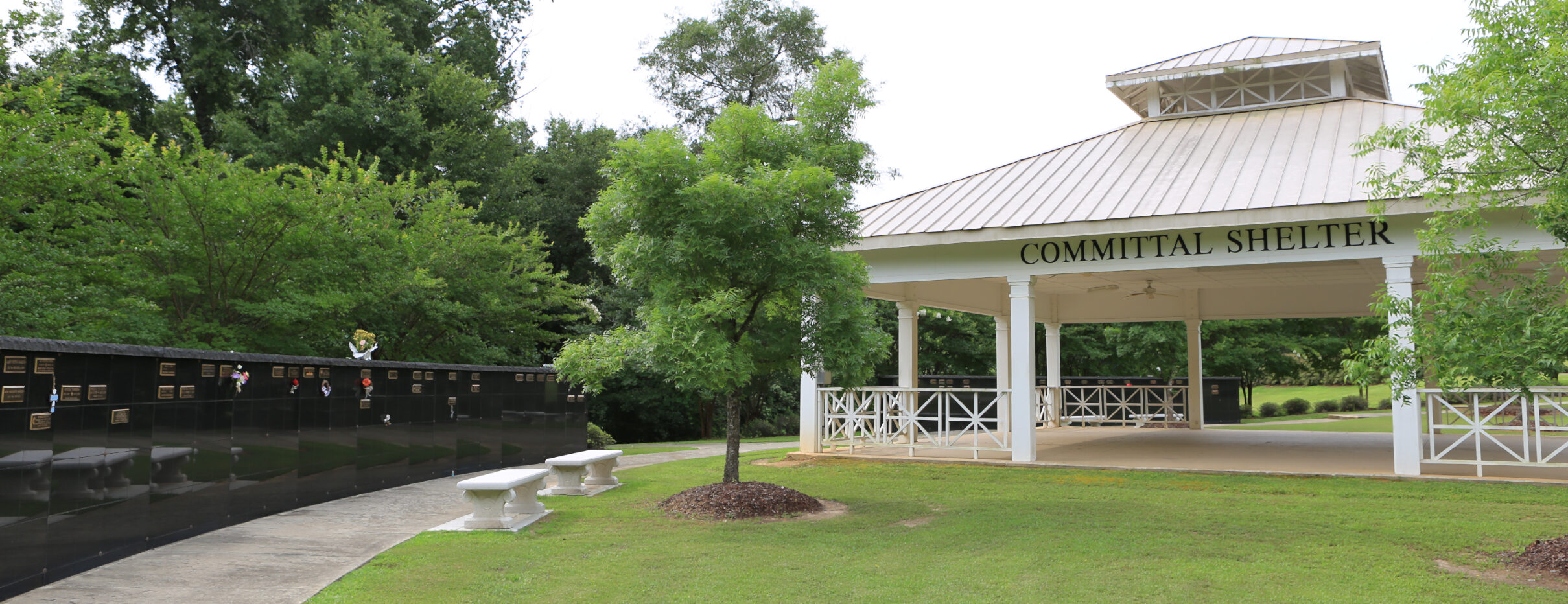 Covered committal shelter with white columns next to a columbarium, benches, and a grassy landscaped area with trees.