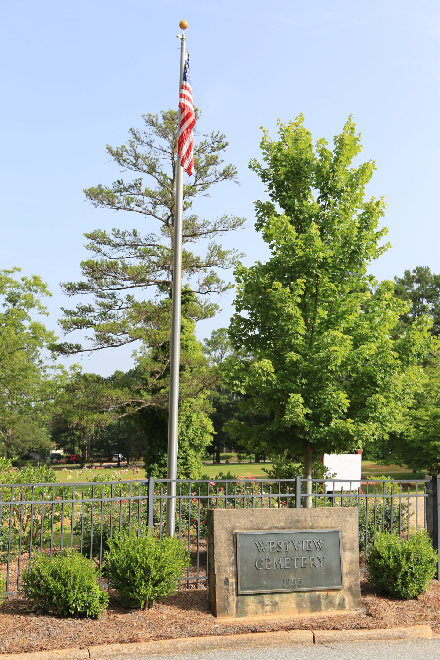 United States flag on a tall flagpole above a stone sign reading "Westview Cemetery," surrounded by landscaped plants and fencing.