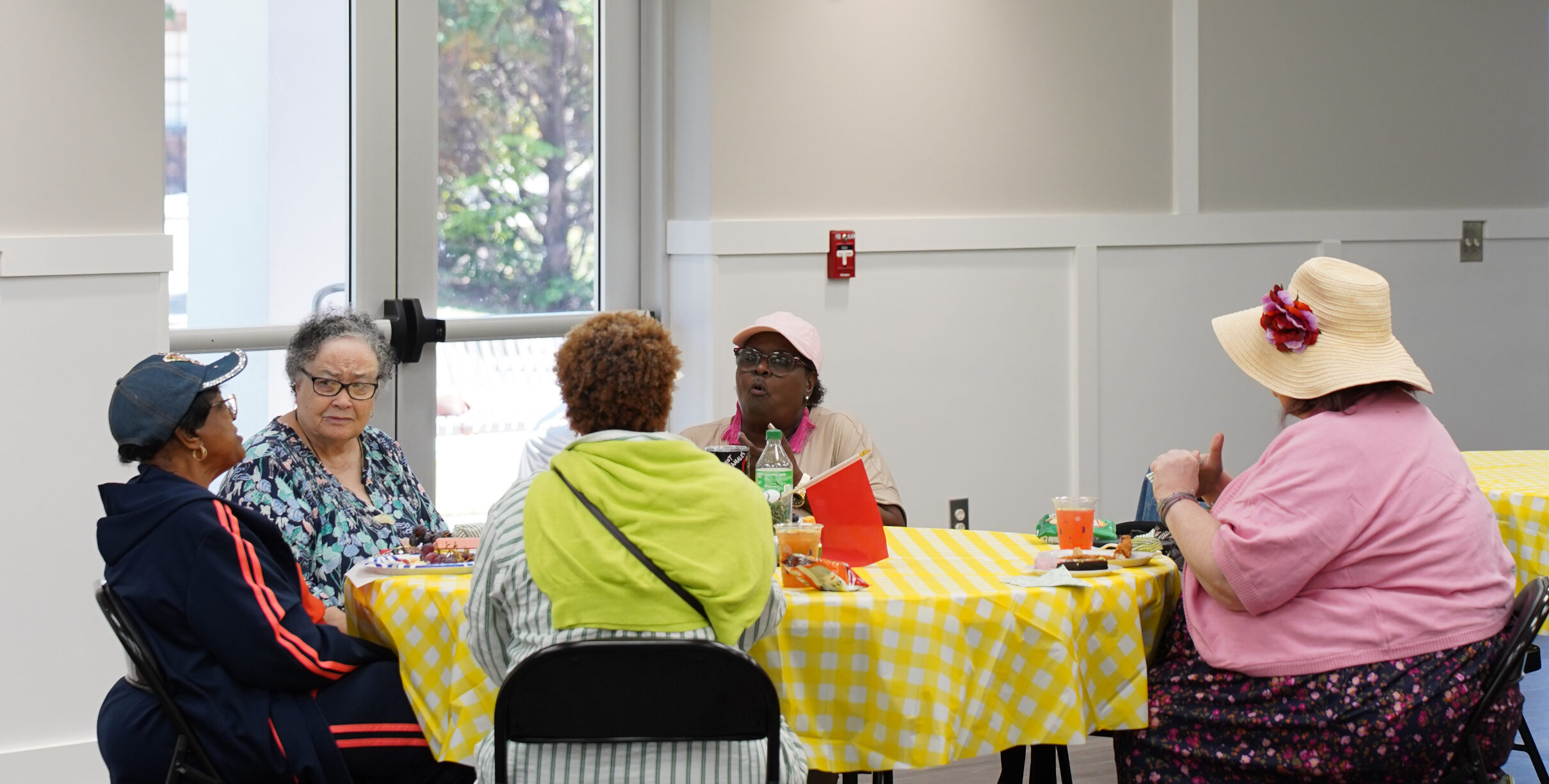 A group of women seated at a round table with a yellow tablecoth, enjoying lunch and engaged in conversation.