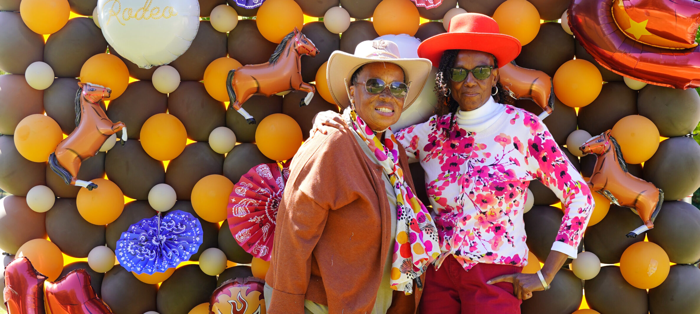 Two adult women standing in front of a colorful, rodeo themed balloon backdrop. 