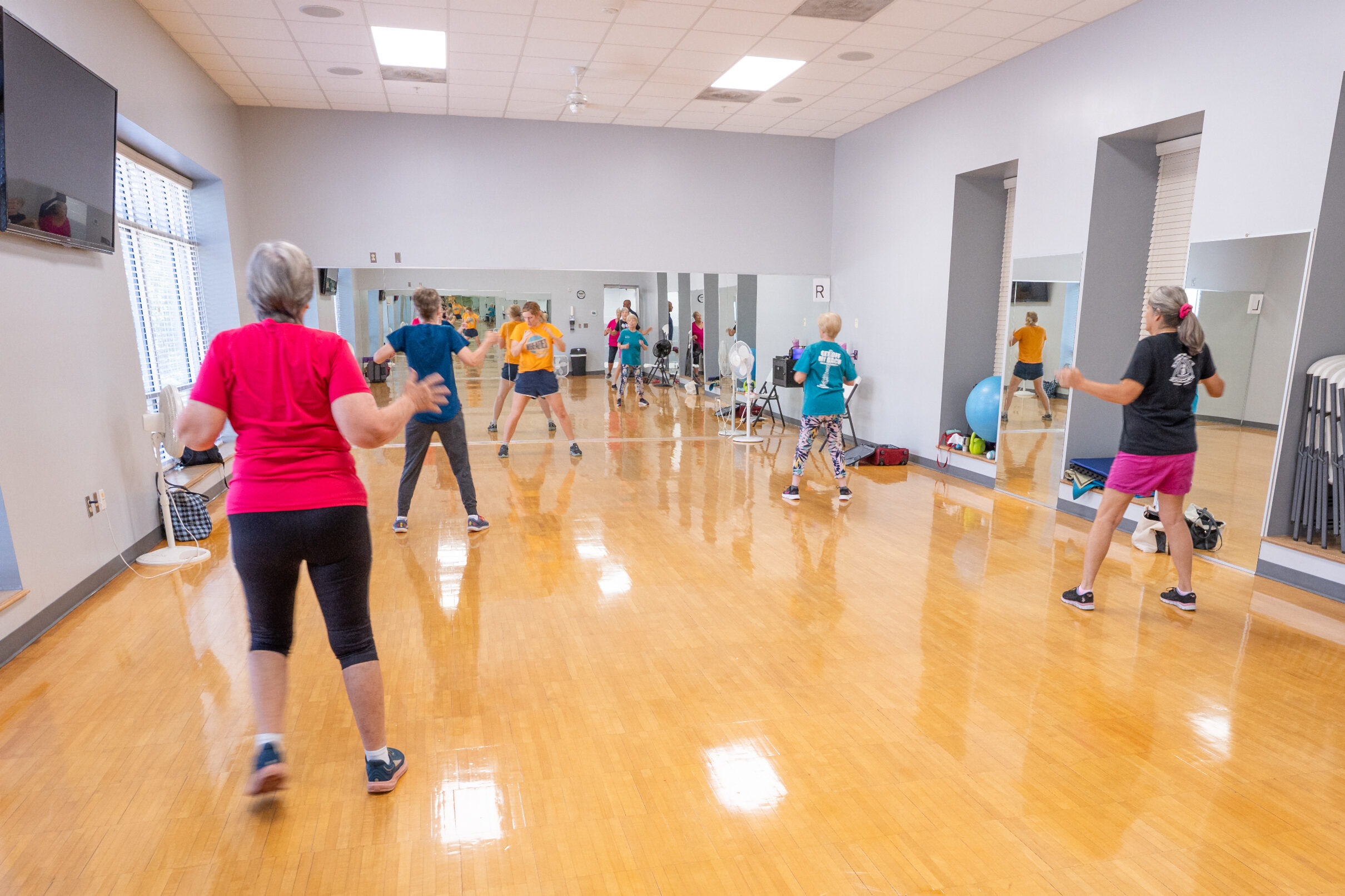 Participants in a group fitness class exercising in a mirrored studio with wood flooring and equipment along the walls.