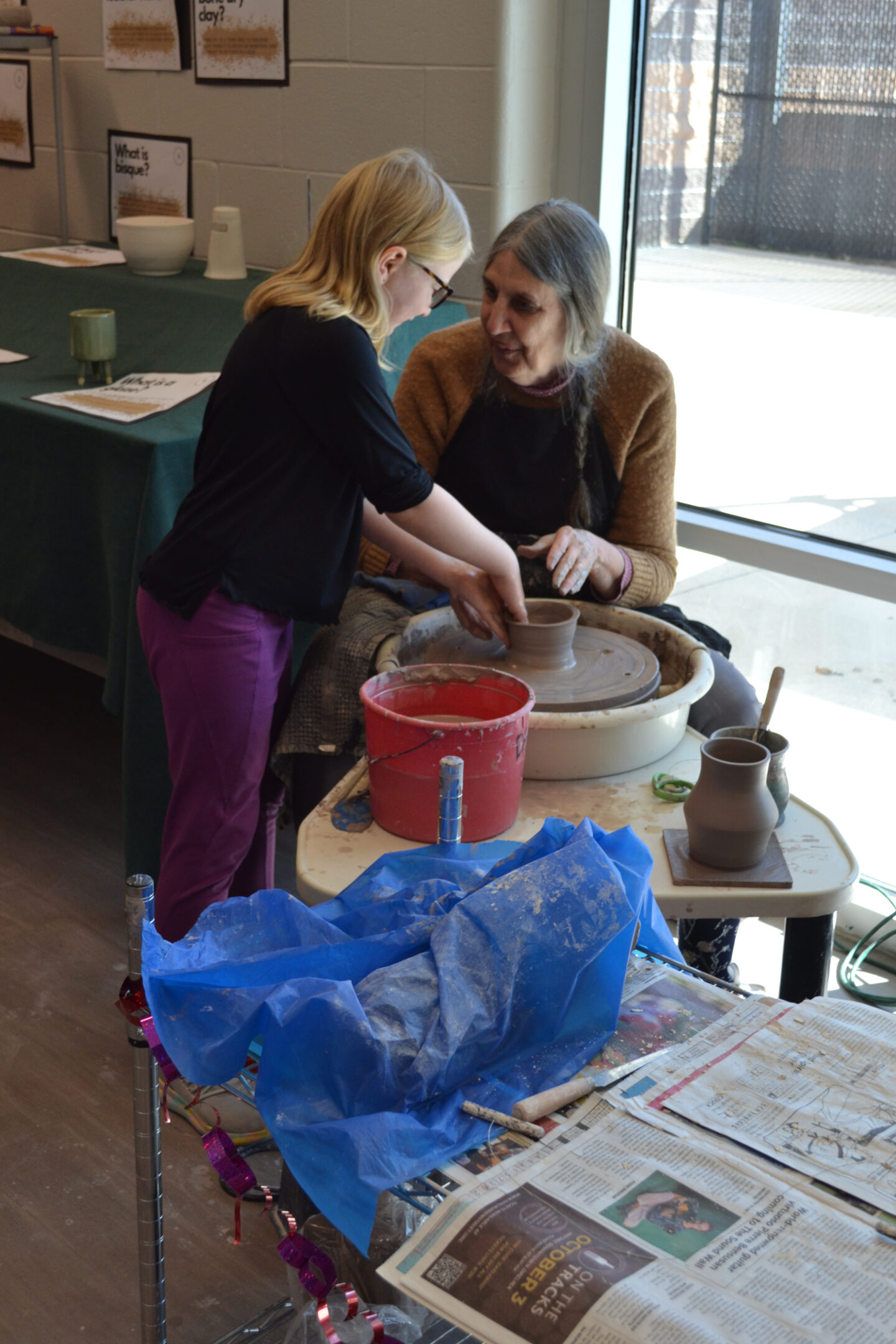 Image shows potters wheel instructor throwing on a wheel and showing a young participant what they are doing.