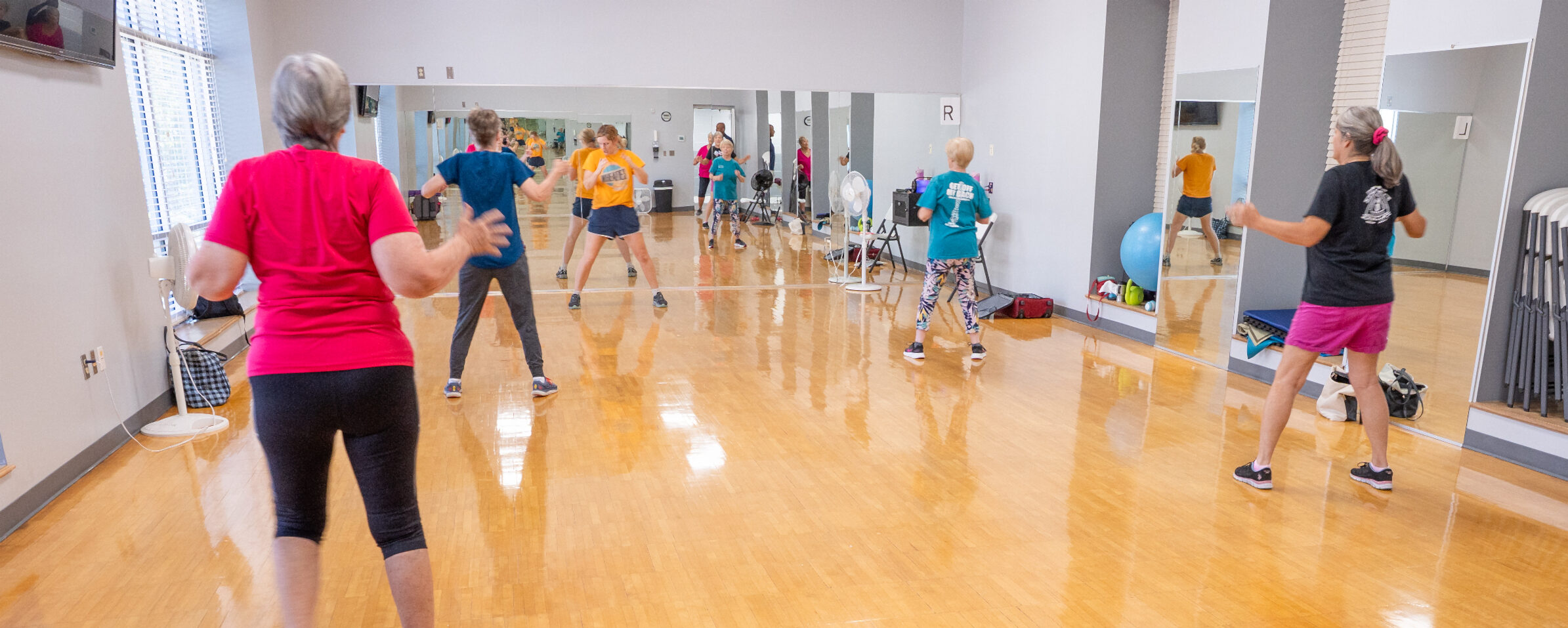 A group of women participating in a fitness class. They are isnide a fitness studio with large mirrors and wooden flooring, dressed in active clothing.