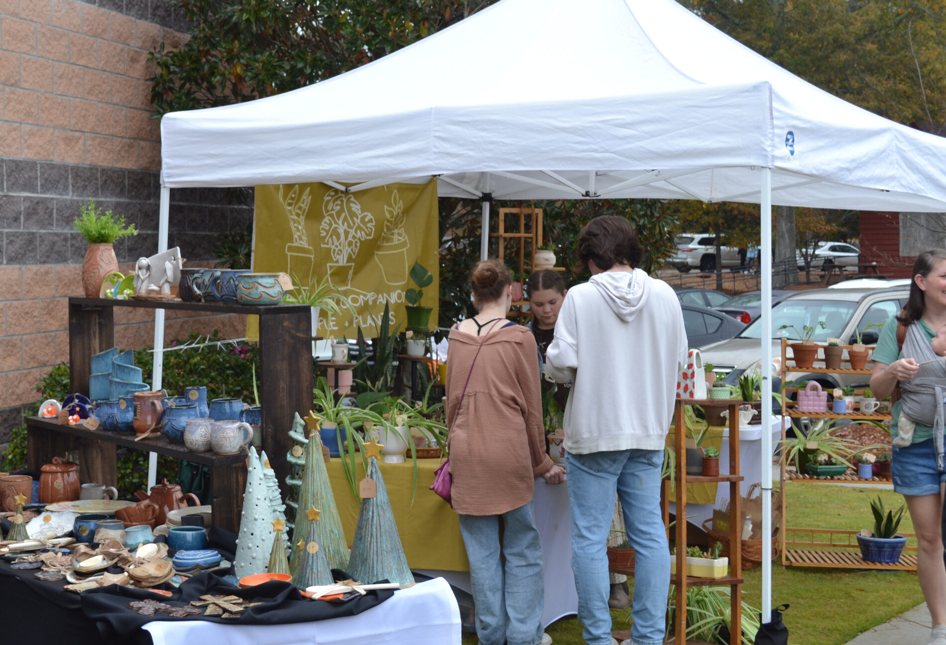 Two people stand with their backs to the viewer at an outdoor booth under a white tent, browsing a display of handmade ceramics, plants, and decorative items arranged on tables and shelves. A vendor stands behind the table, and another person views the booth as they walk past.