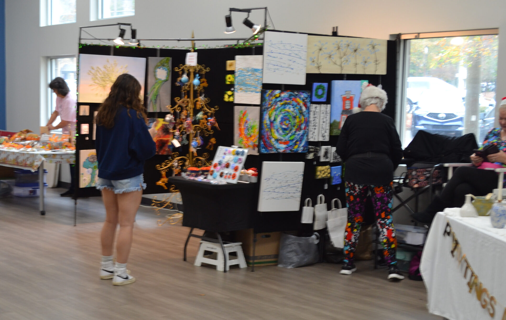 Two people standing in the foreground of an indoor booth at the Holiday Art Sale, facing tables and dsiplay panels filled with colorful paintings and handmade items. Additional tables and a person browsing are visible in the background.